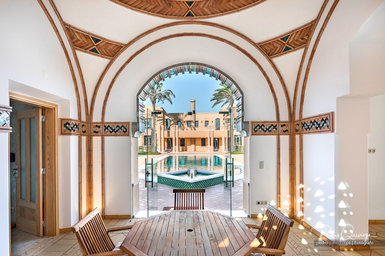 Arco em madeira emoldura vista para piscina e edifício de estilo árabe com palmeiras sob céu azul