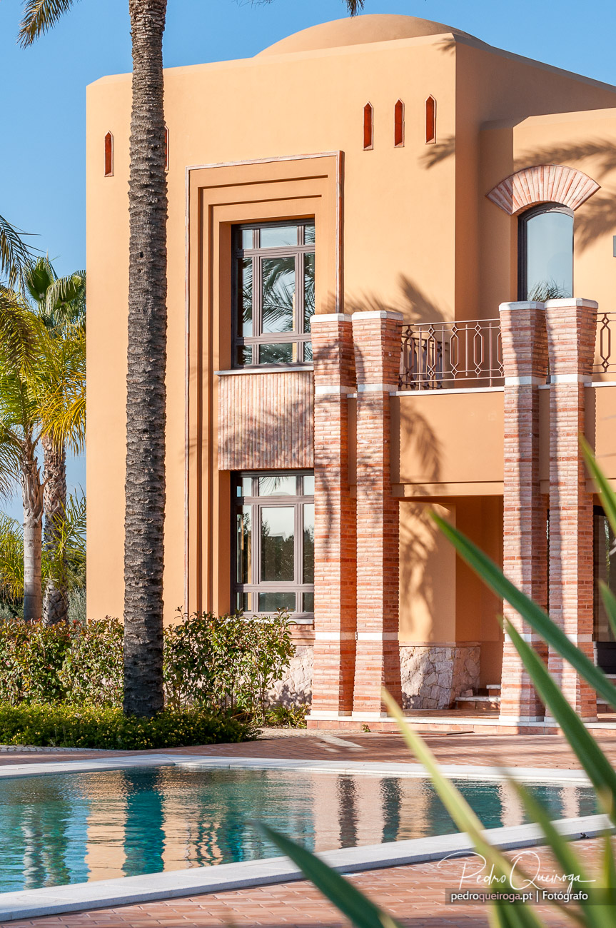 Moradia de tons bege com detalhes em tijolo, varanda e janelas amplas, rodeada por palmeiras e piscina ao sol.