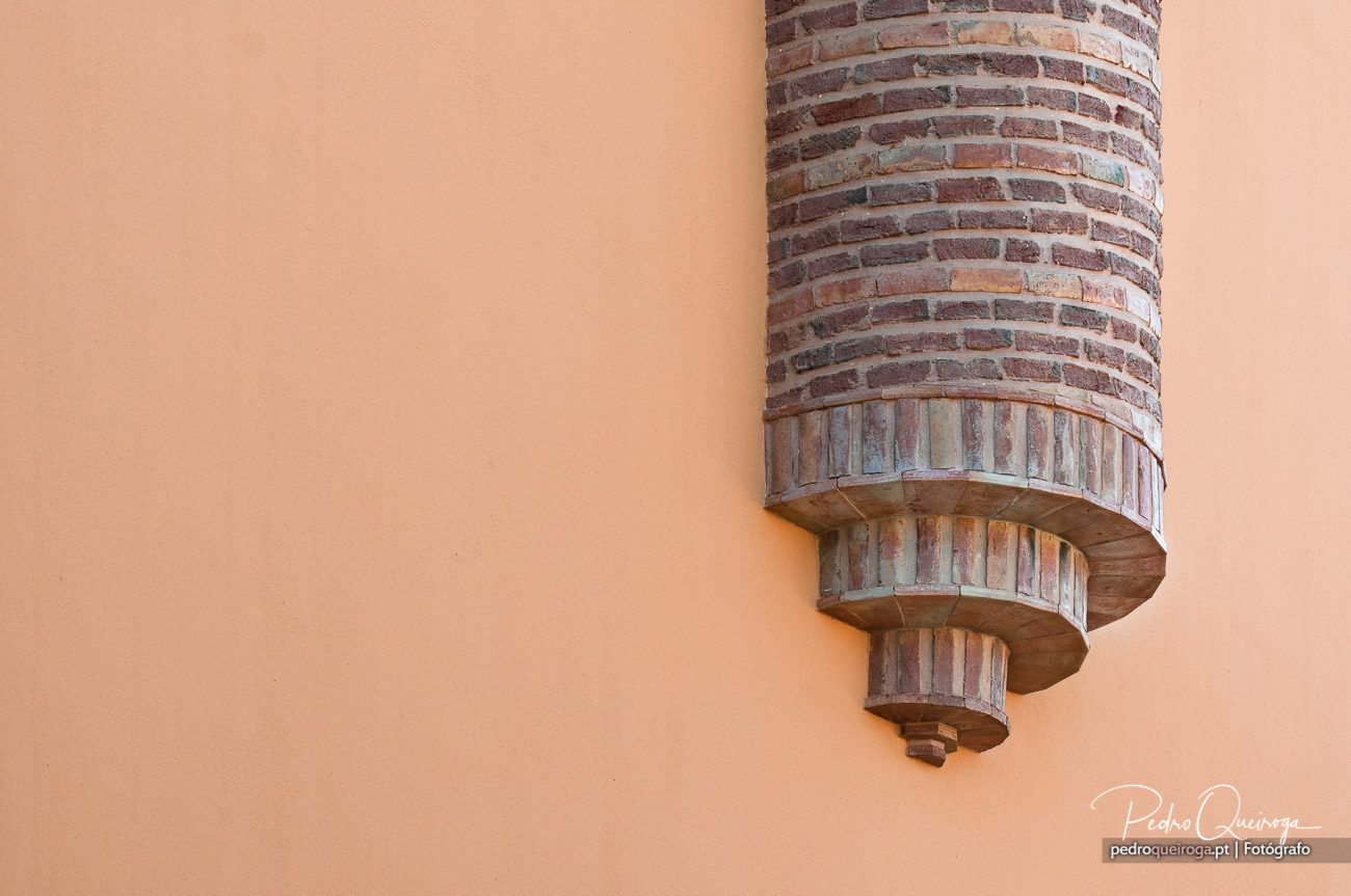Coluna cilíndrica em tijolo vermelho com detalhes arquitetónicos em relevo sobre parede cor de pêssego suave