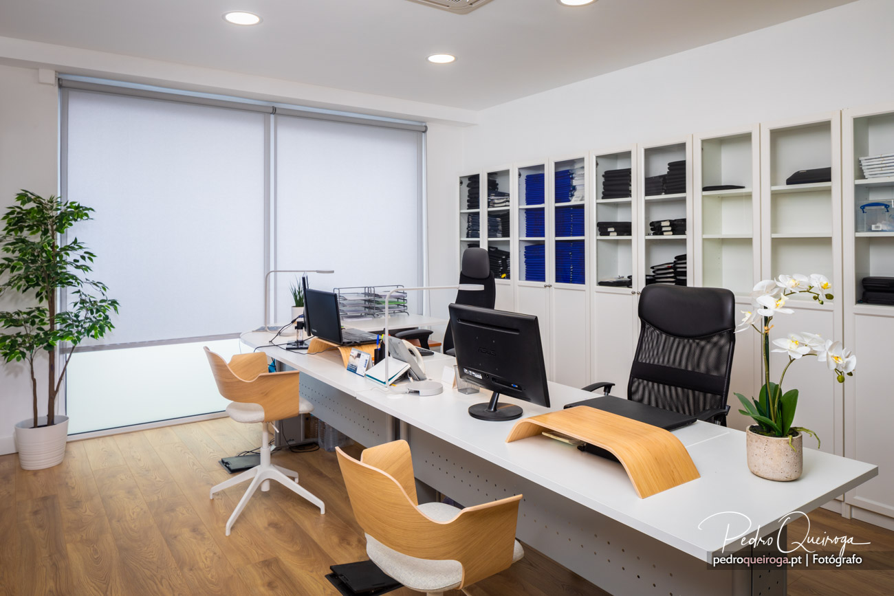 Escritório moderno com secretárias brancas, cadeiras ergonómicas, estantes organizadas e luz natural abundante através de persianas.