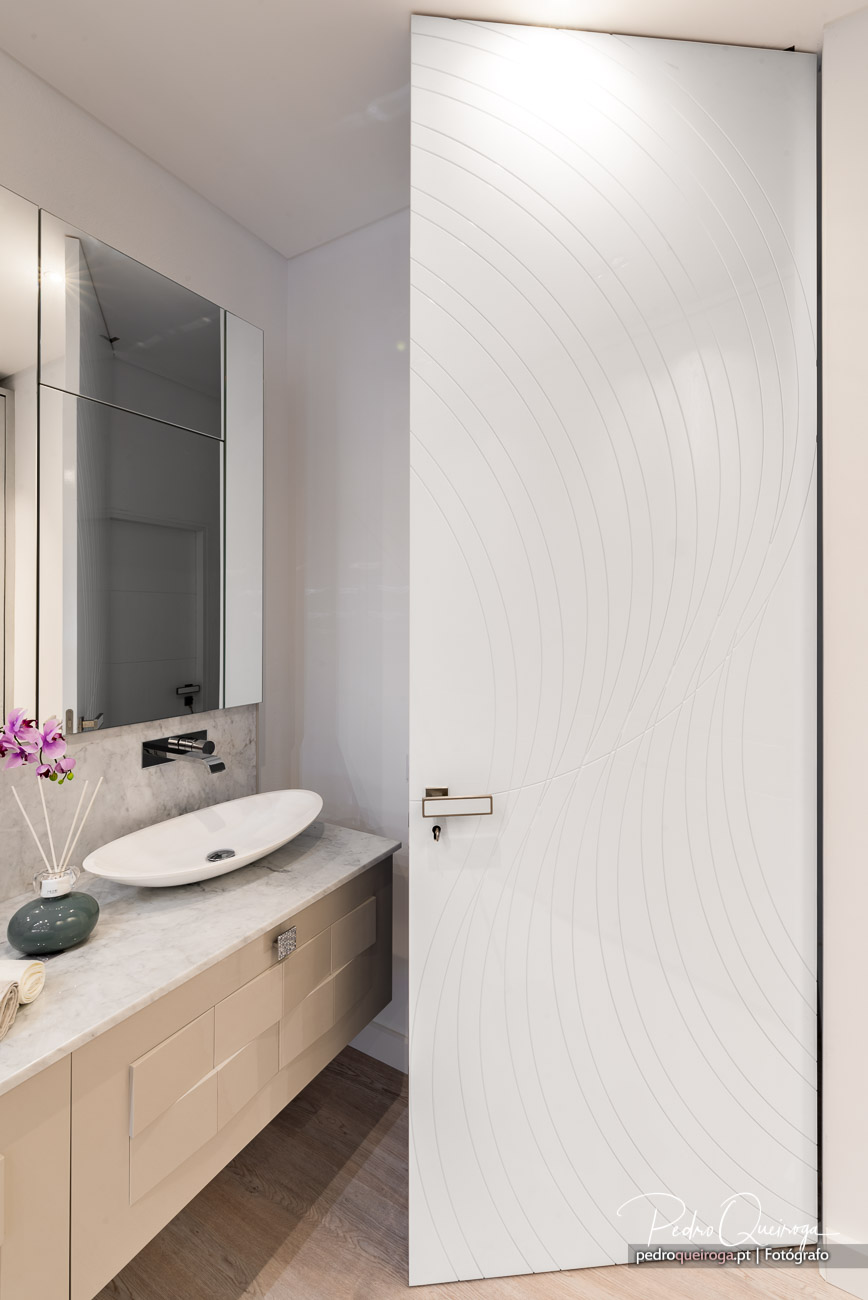 Casa de banho moderna com porta branca texturada, bancada em pedra clara e espelho elegante sobre o lavatório oval.