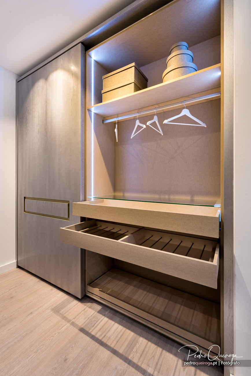 Roupeiro moderno em madeira clara com iluminação LED, cabides brancos, prateleiras superiores e gavetas abertas organizadas