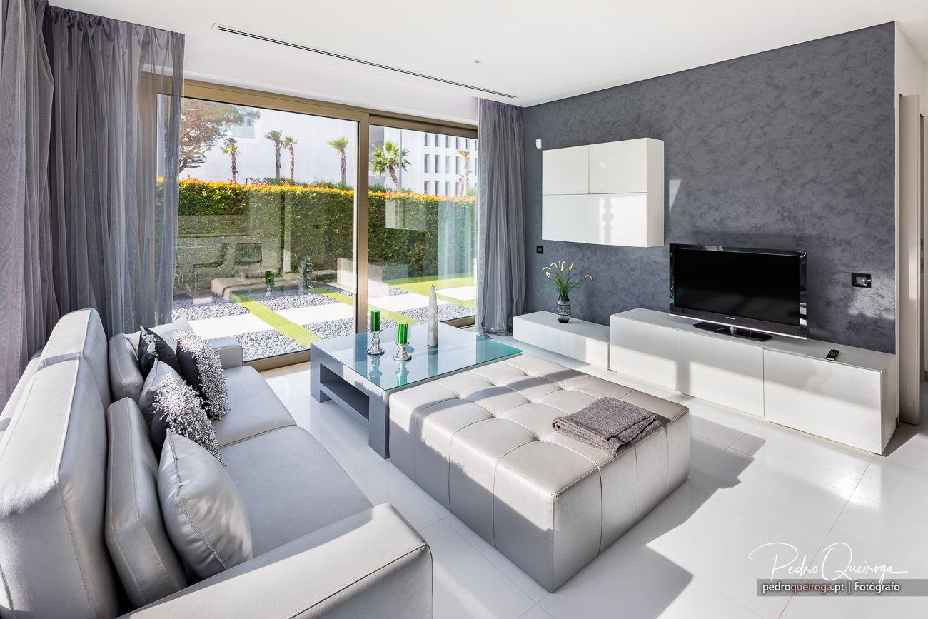 Sala moderna com sofás cinzentos, parede texturizada escura, móveis brancos e ampla janela com vista para jardim e palmeiras
