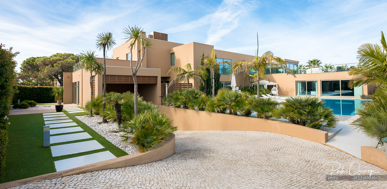 Moradia moderna em tons bege com jardim tropical, piscina azul e calçada em calçada portuguesa sob céu azul claro