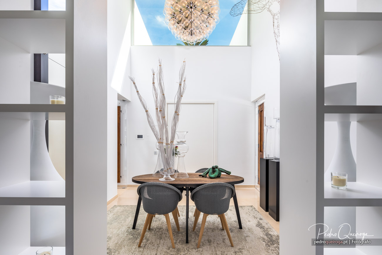 Sala de jantar moderna com mesa redonda, cadeiras cinzentas, candeeiro esférico e pé-direito duplo com luz natural.