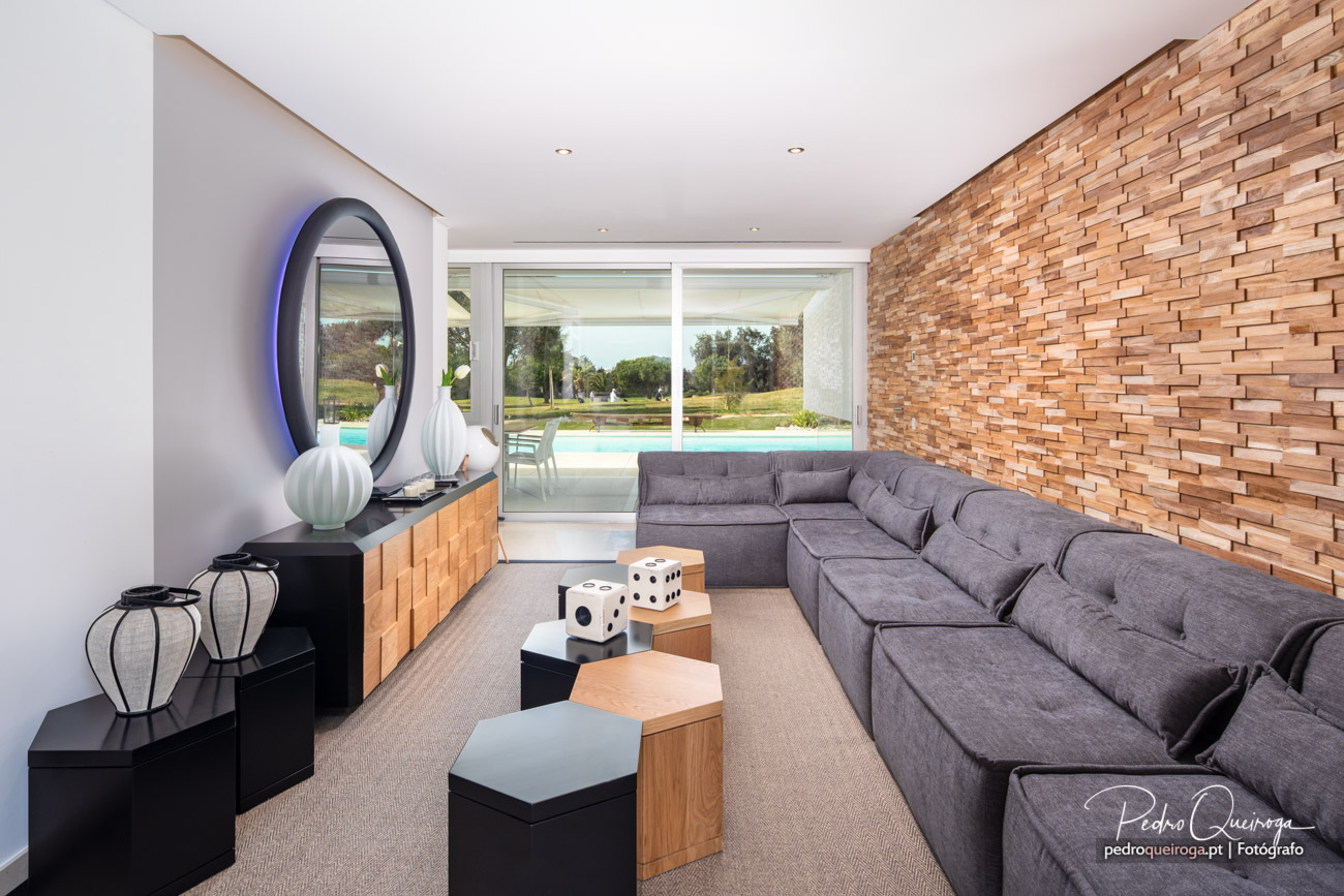 Sala moderna com sofá cinzento, parede em madeira texturizada e vista para piscina através de amplas janelas envidraçadas