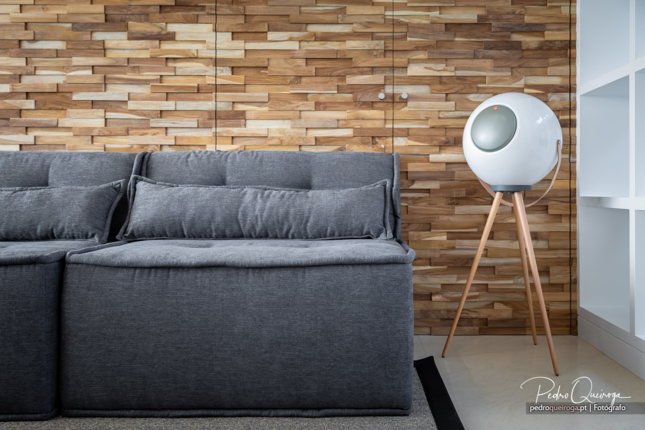 Sofá cinzento confortável junto a parede revestida com ripas de madeira e escultura branca moderna com tripé