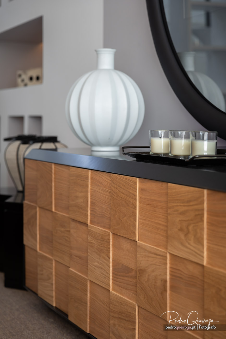 Aparador em madeira com tampo preto, decorado com jarro branco canelado, velas e espelho redondo ao fundo.