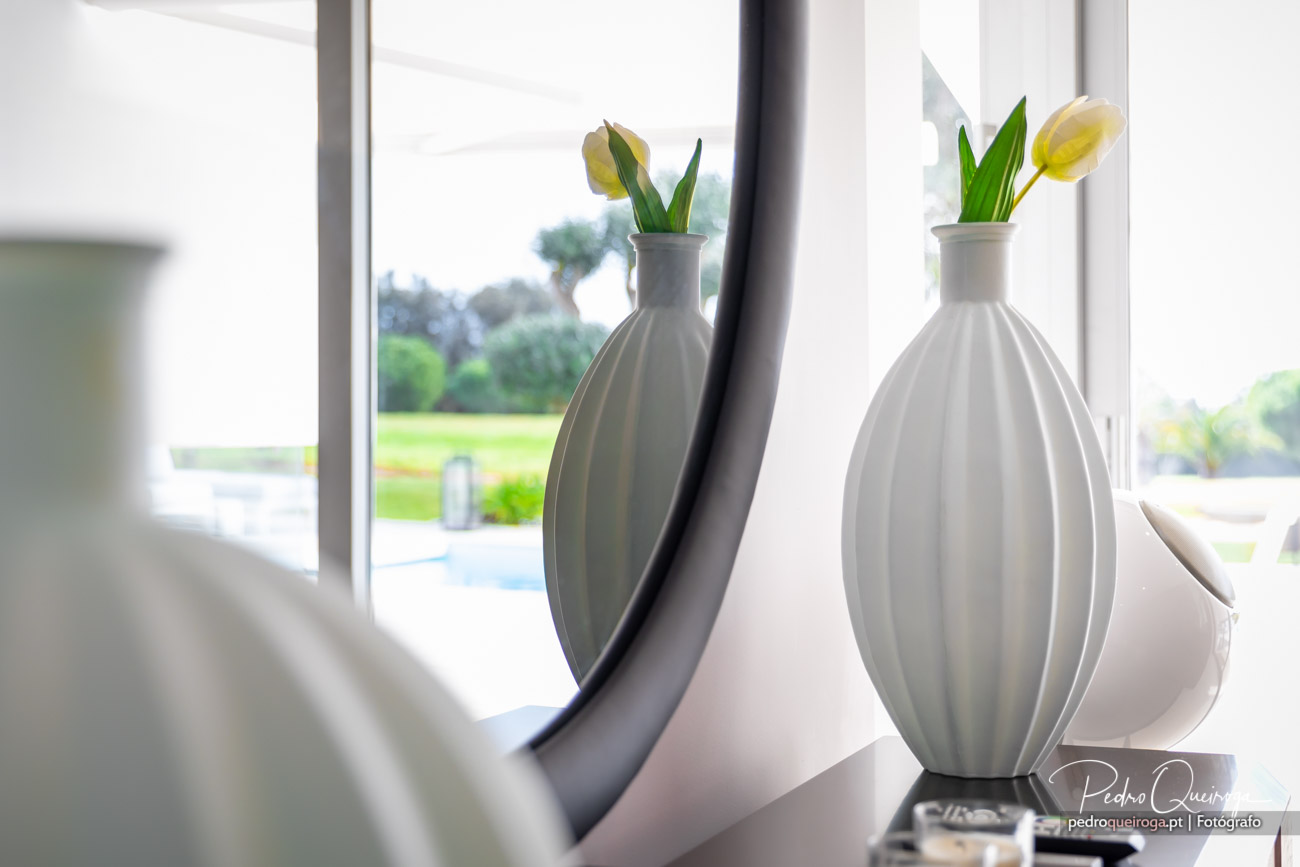 Jarras brancas texturizadas com flores amarelas sobre prateleira junto a janela ampla com vista para jardim exterior