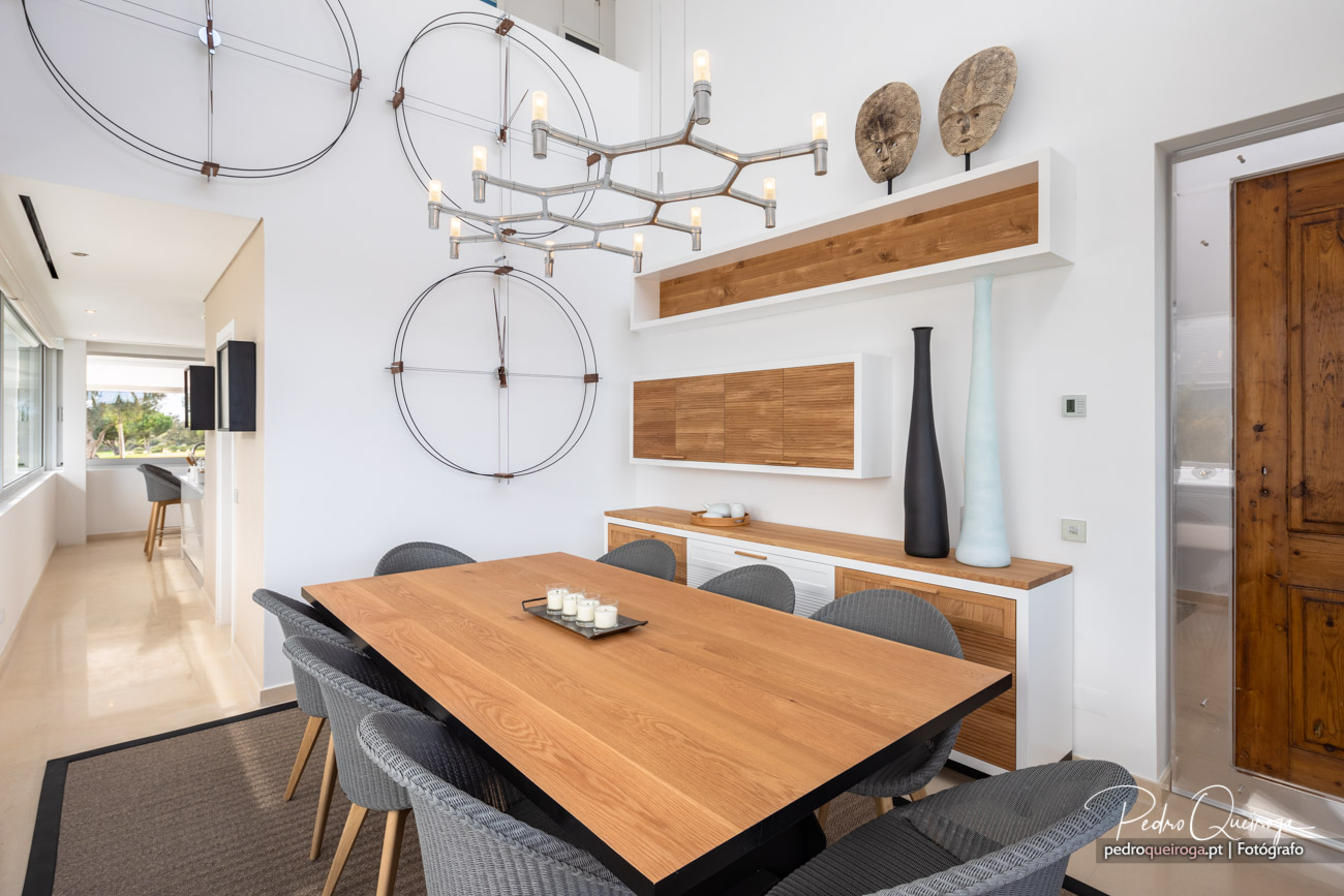 Sala de jantar moderna com mesa em madeira, cadeiras cinzas, lustre geométrico e móvel branco com detalhes em madeira.