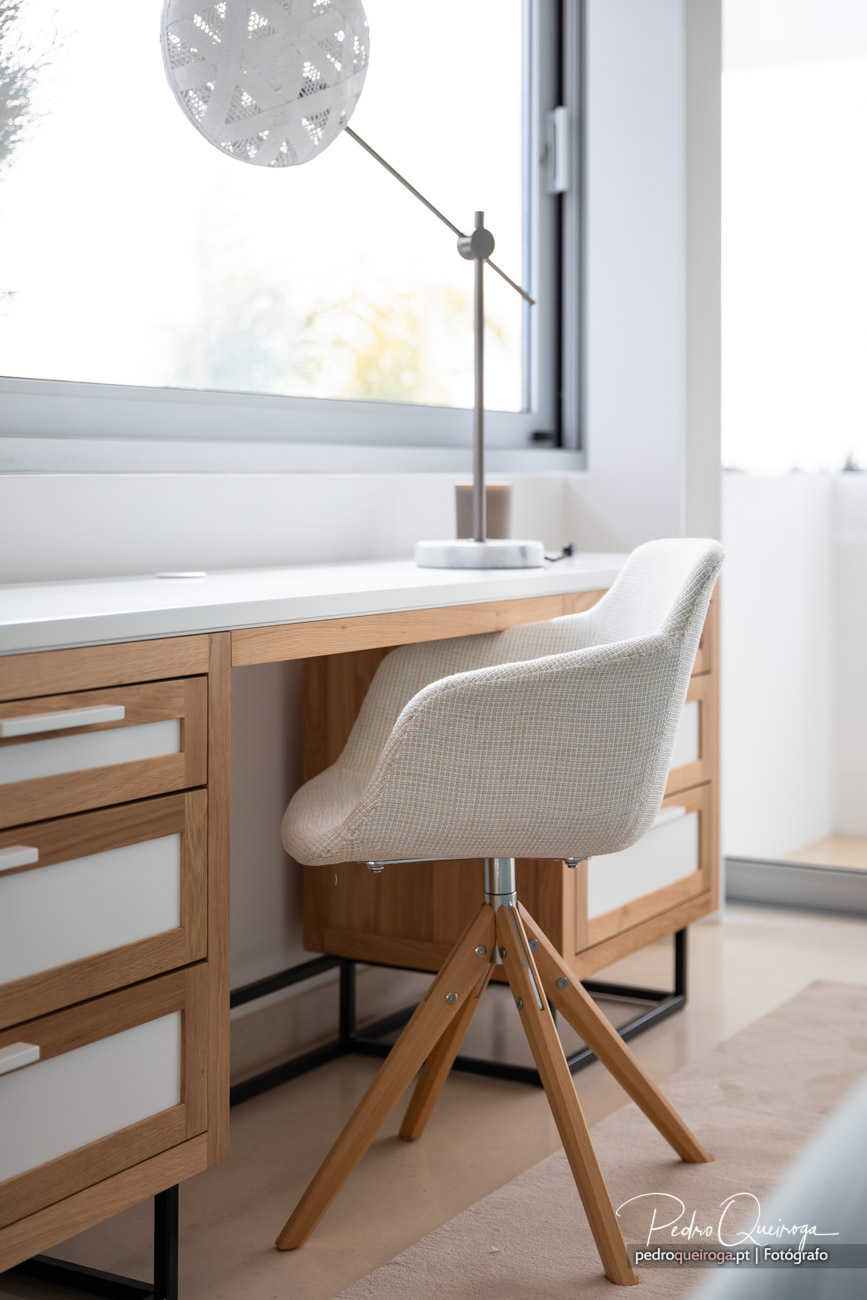 Cadeira bege estofada com pés em madeira junto a secretária minimalista com candeeiro decorativo e janela luminosa