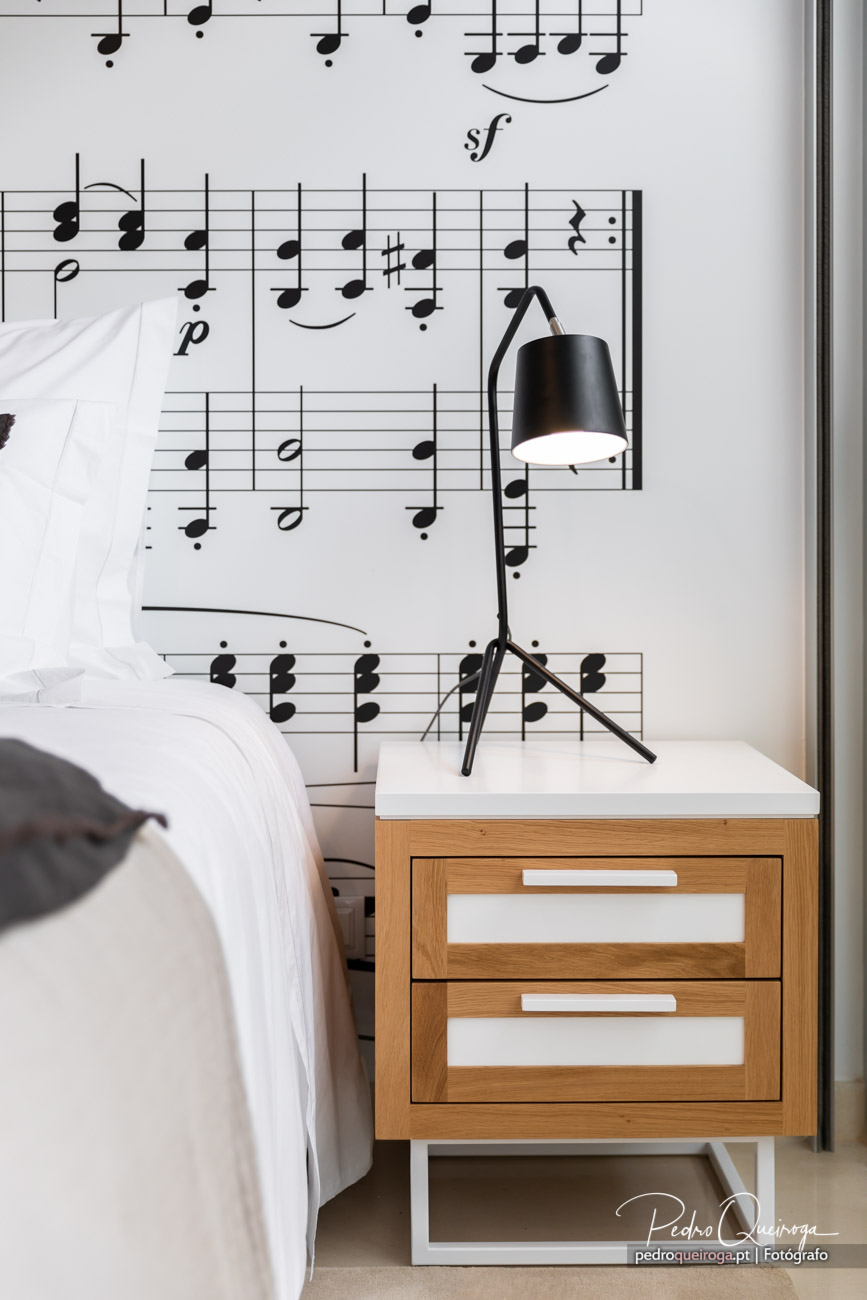 Quarto com mesinha de cabeceira em madeira, candeeiro preto moderno e parede com partitura musical como decoração