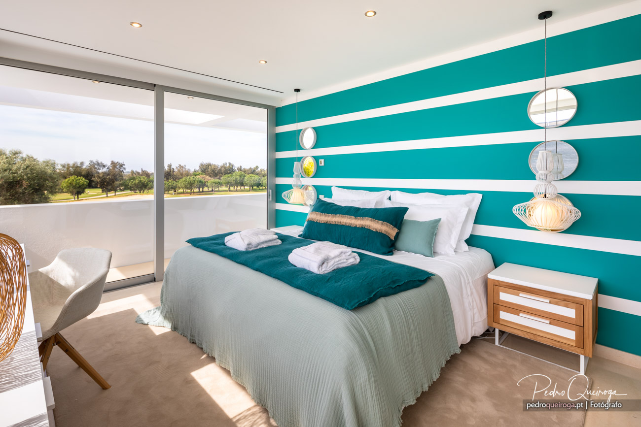 Quarto moderno com parede turquesa às riscas brancas, cama ampla e grandes janelas com vista para campo verde