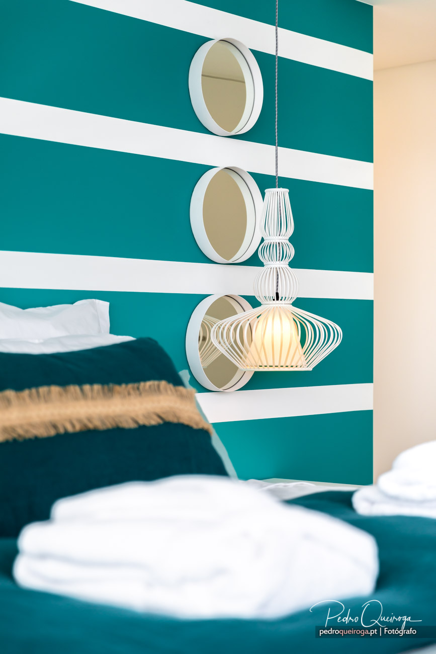 Quarto moderno com parede turquesa às riscas brancas, espelhos redondos, candeeiro suspenso e roupa de cama branca