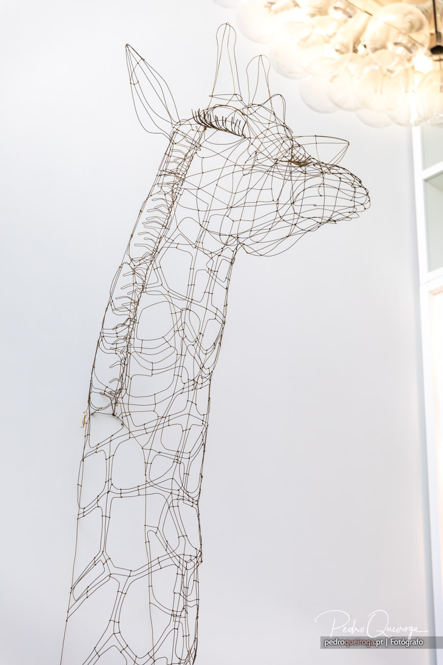 Escultura de girafa em arame metálico com traçado geométrico junto a parede branca e candeeiro dourado ao fundo