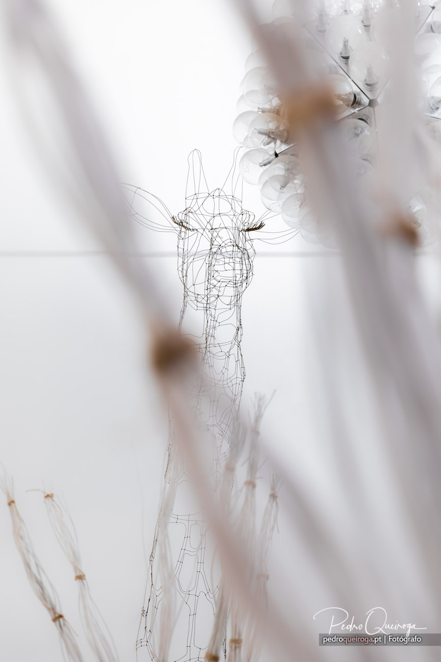 Escultura minimalista em arame formando uma girafa suspensa, com lustre desfocado ao fundo em tons neutros e suaves.