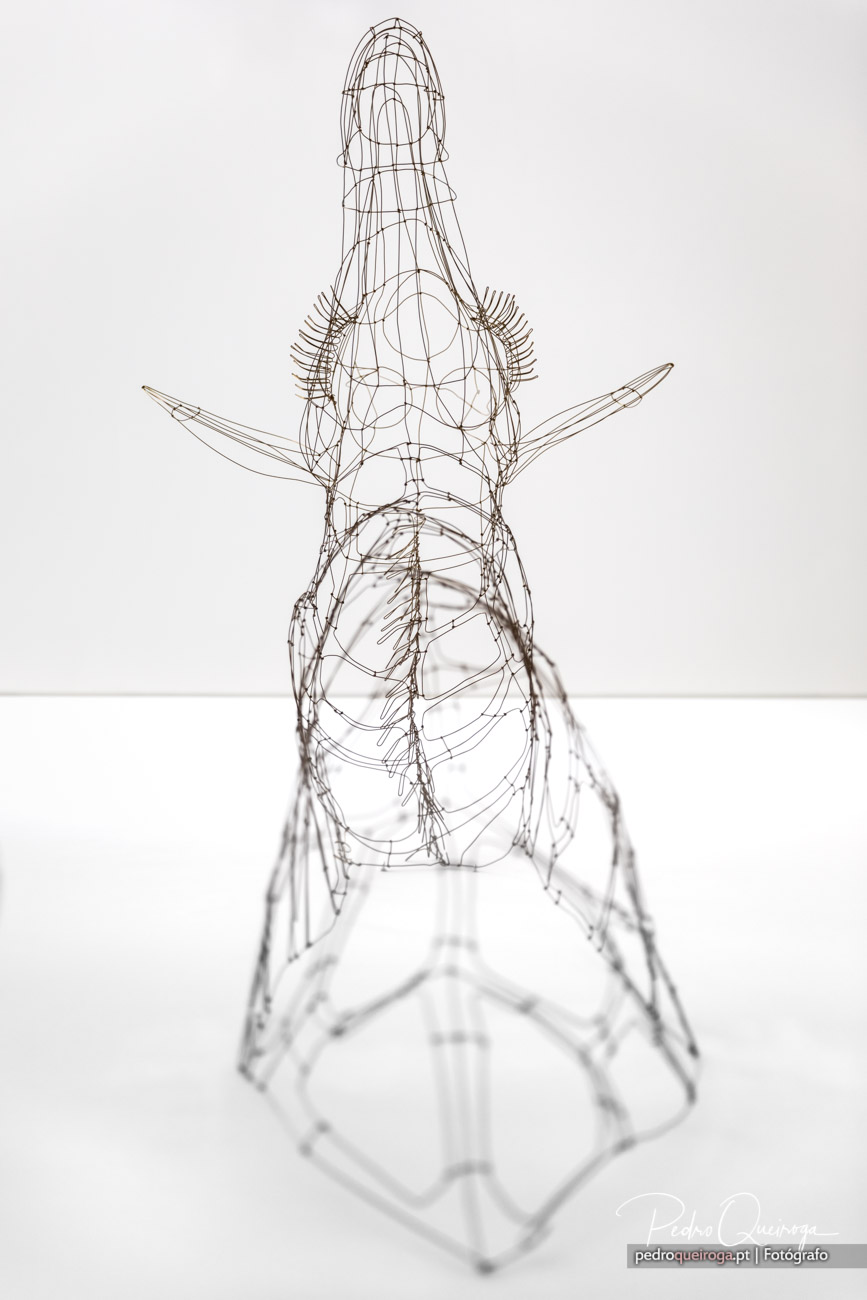 Escultura de arame em forma de figura feminina ajoelhada com braços abertos, sobre fundo branco minimalista