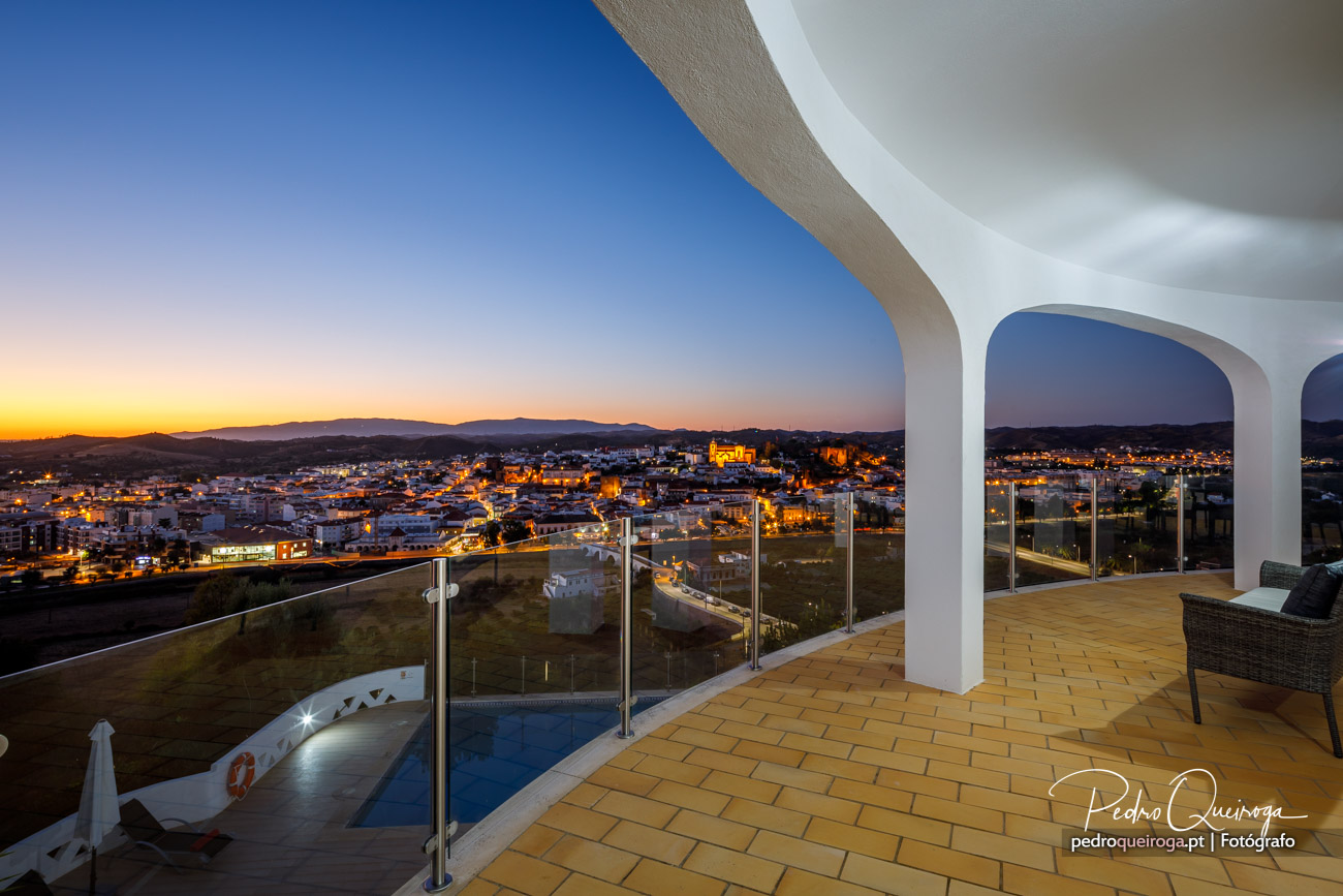 Terraço moderno com piscina e arcos brancos ao entardecer, com vista panorâmica sobre cidade iluminada e colinas ao fundo