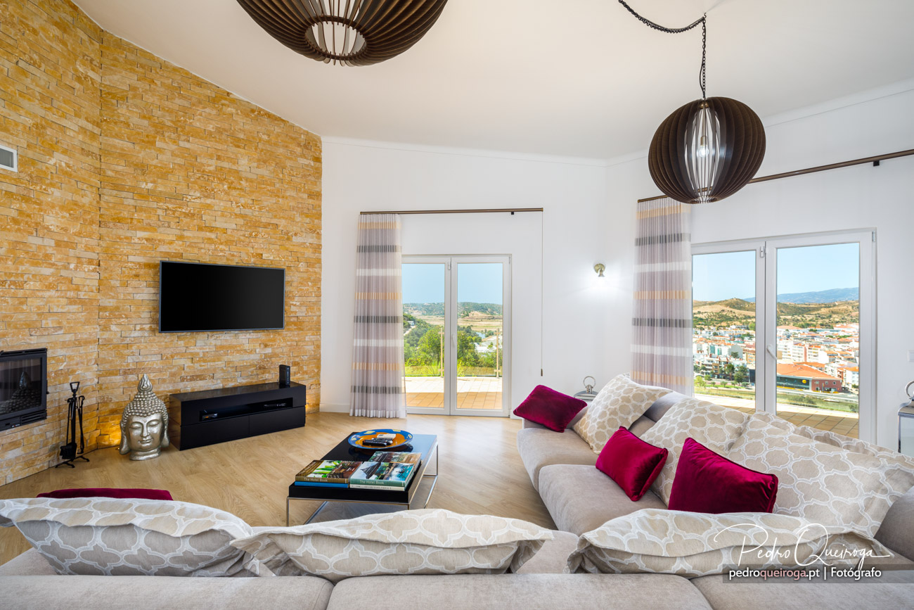 Sala ampla com parede em pedra amarela, sofá bege com almofadas bordeaux e janelas panorâmicas com vista para o exterior