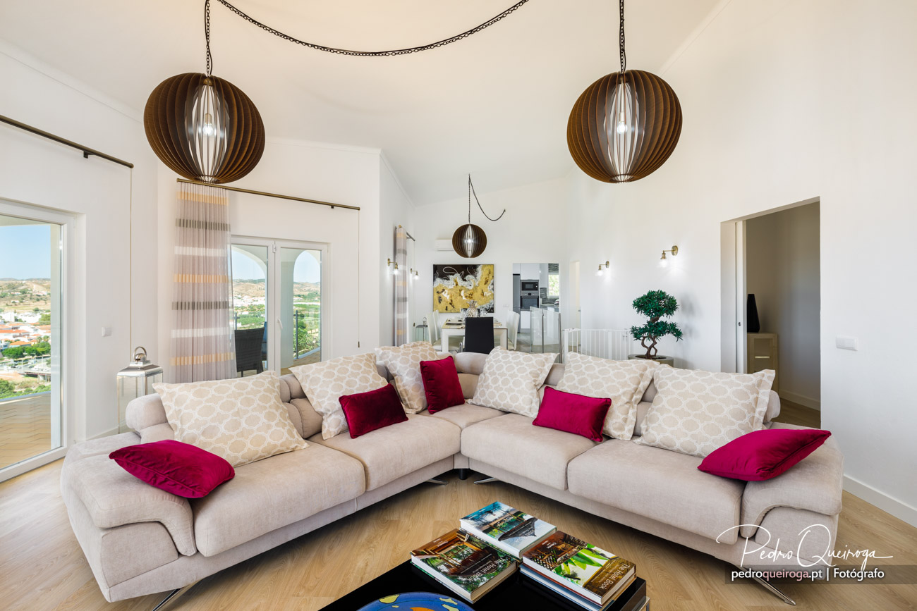 Sala ampla e luminosa com sofá bege, almofadas vermelhas e candeeiros suspensos em madeira num imóvel moderno