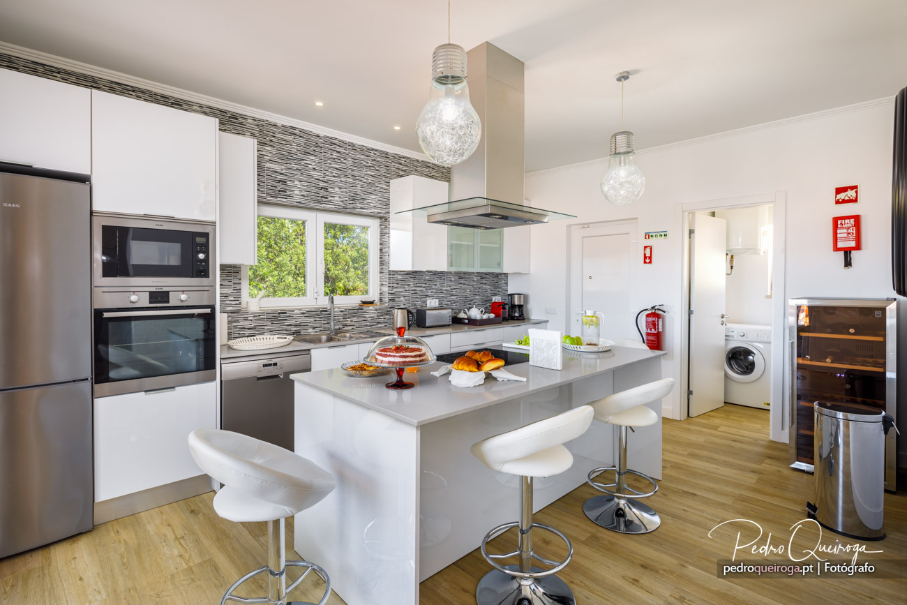 Cozinha moderna com ilha central, bancadas cinza, armários brancos, eletrodomésticos embutidos e pavimento em madeira clara