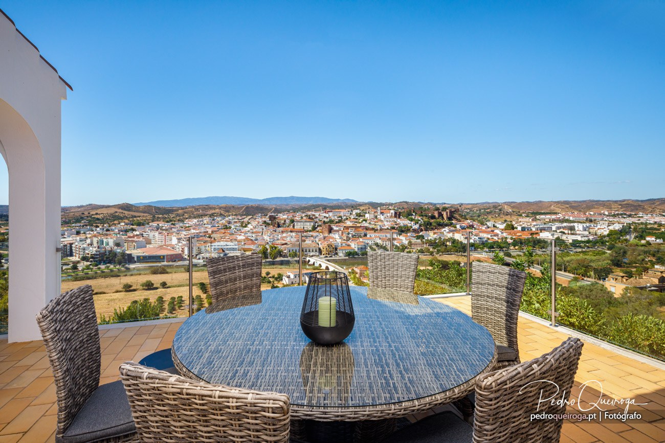 Terraço com mesa redonda em rattan e cadeiras, vista panorâmica sobre a cidade e colinas ao fundo sob céu azul.