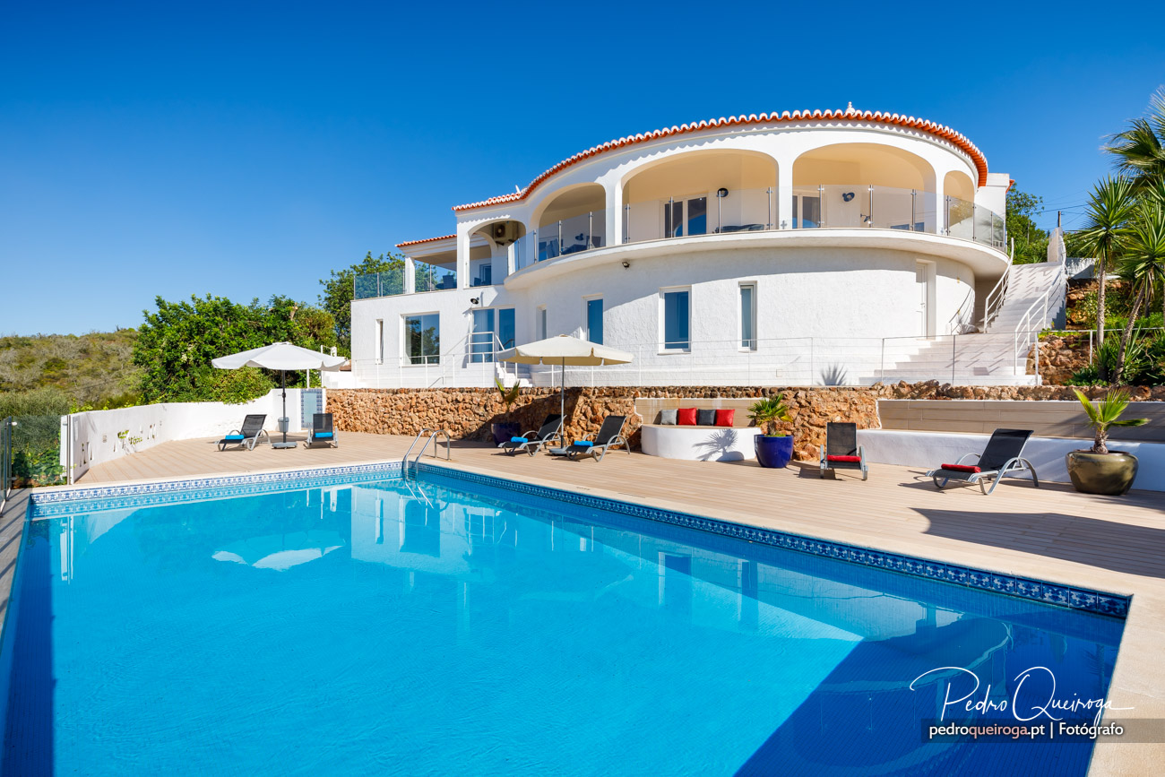 Moradia moderna de arquitetura curva com telhado de telha, piscina azul e espreguiçadeiras sob céu límpido