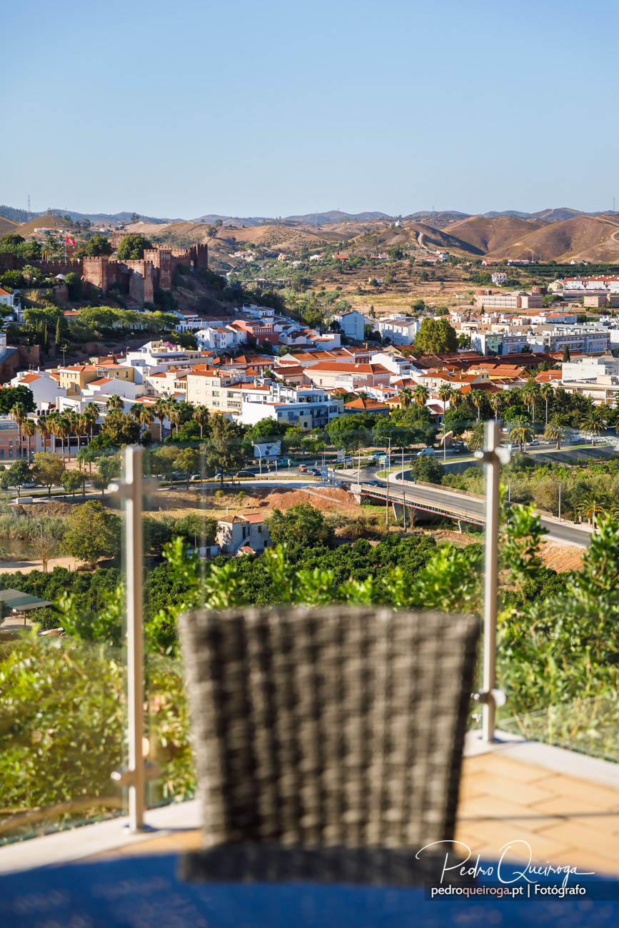 Vista panorâmica de vila alentejana com castelo, casas de telhado laranja e colinas ao fundo, vista de um terraço com cadeira