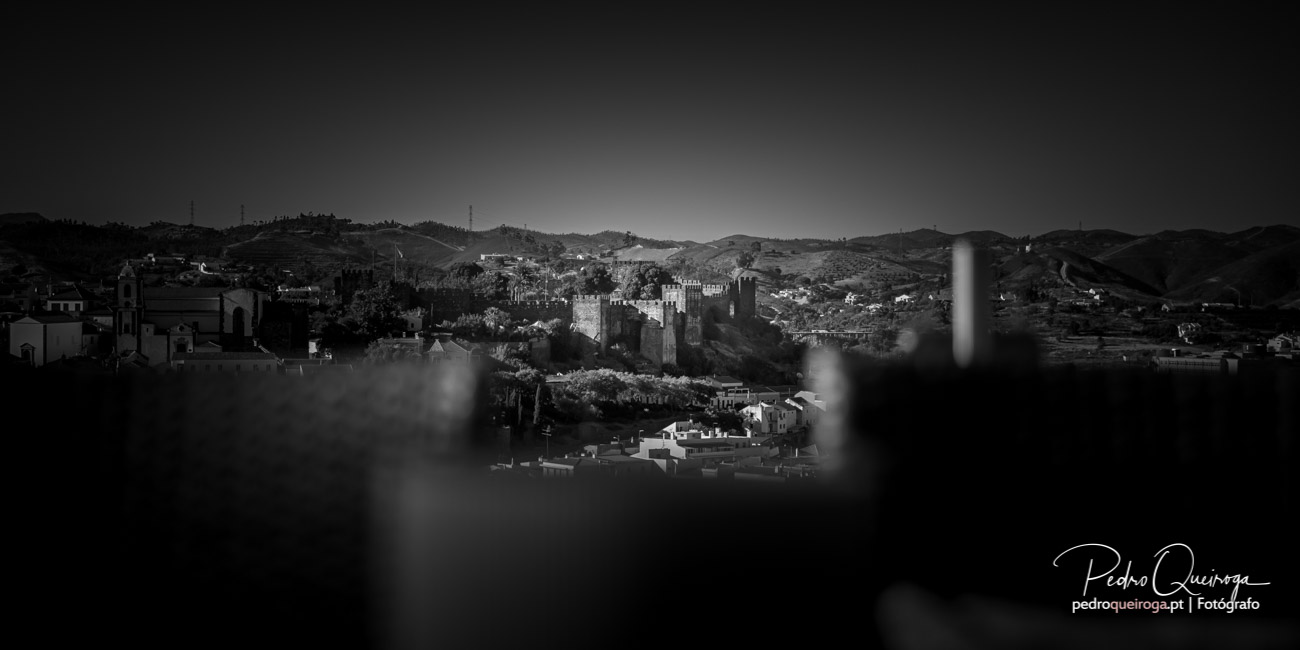 Castelo medieval envolto em névoa sobre povoação, com colinas onduladas ao fundo e igreja à esquerda a preto e branco