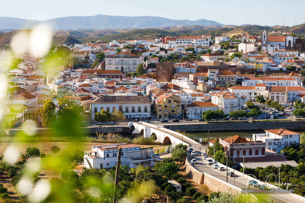 Vista panorâmica de vila alentejana com casas brancas e telhados cor de telha, ponte antiga sobre o rio e serras ao fundo