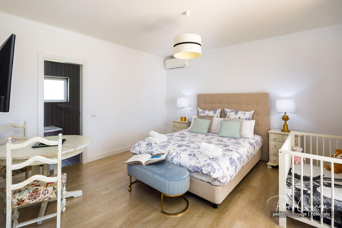 Quarto luminoso com cama de casal, mesitas, berço branco e casa de banho privativa captado por fotógrafo profissional