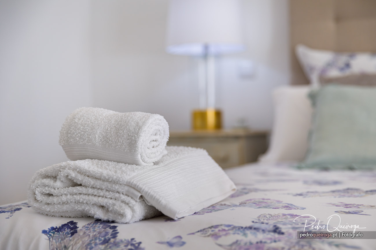 Toalhas brancas dobradas sobre cama com roupa florida, difusor aromático ao fundo num ambiente de quarto acolhedor