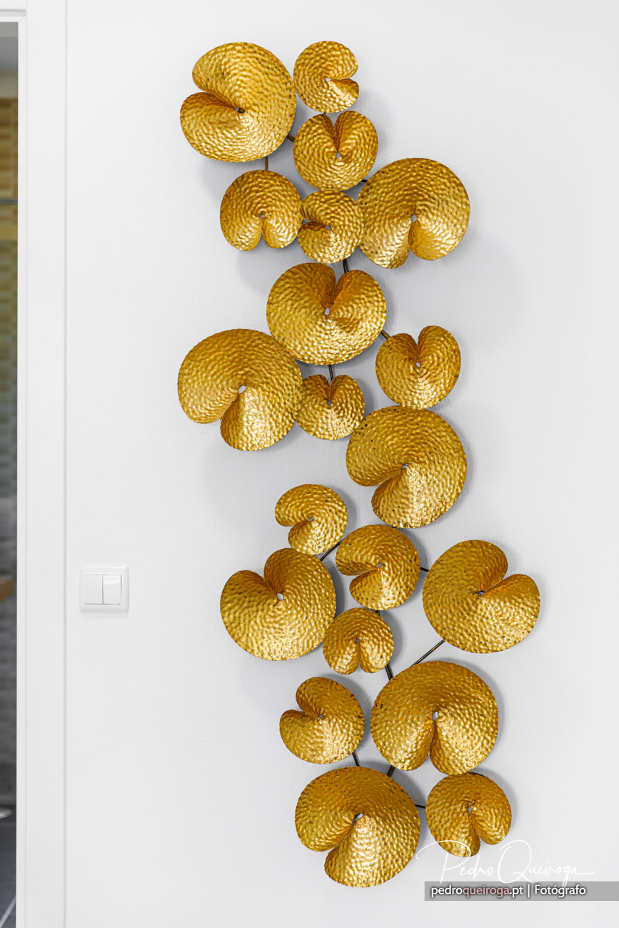 Escultura decorativa vertical em parede branca com elementos dourados texturizados dispostos organicamente