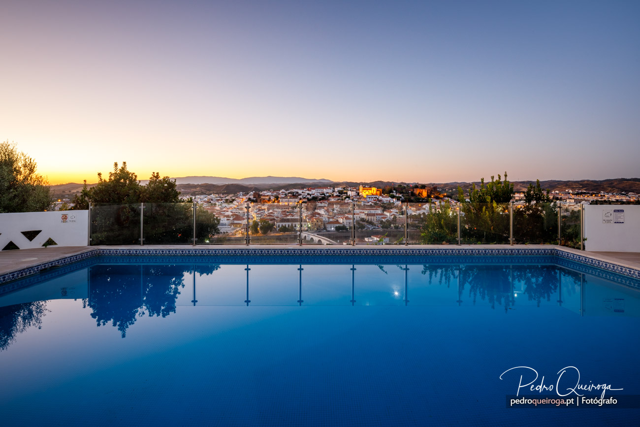 Piscina tranquila ao entardecer com vista panorâmica sobre vila algarvia e colinas ao fundo sob céu crepuscular