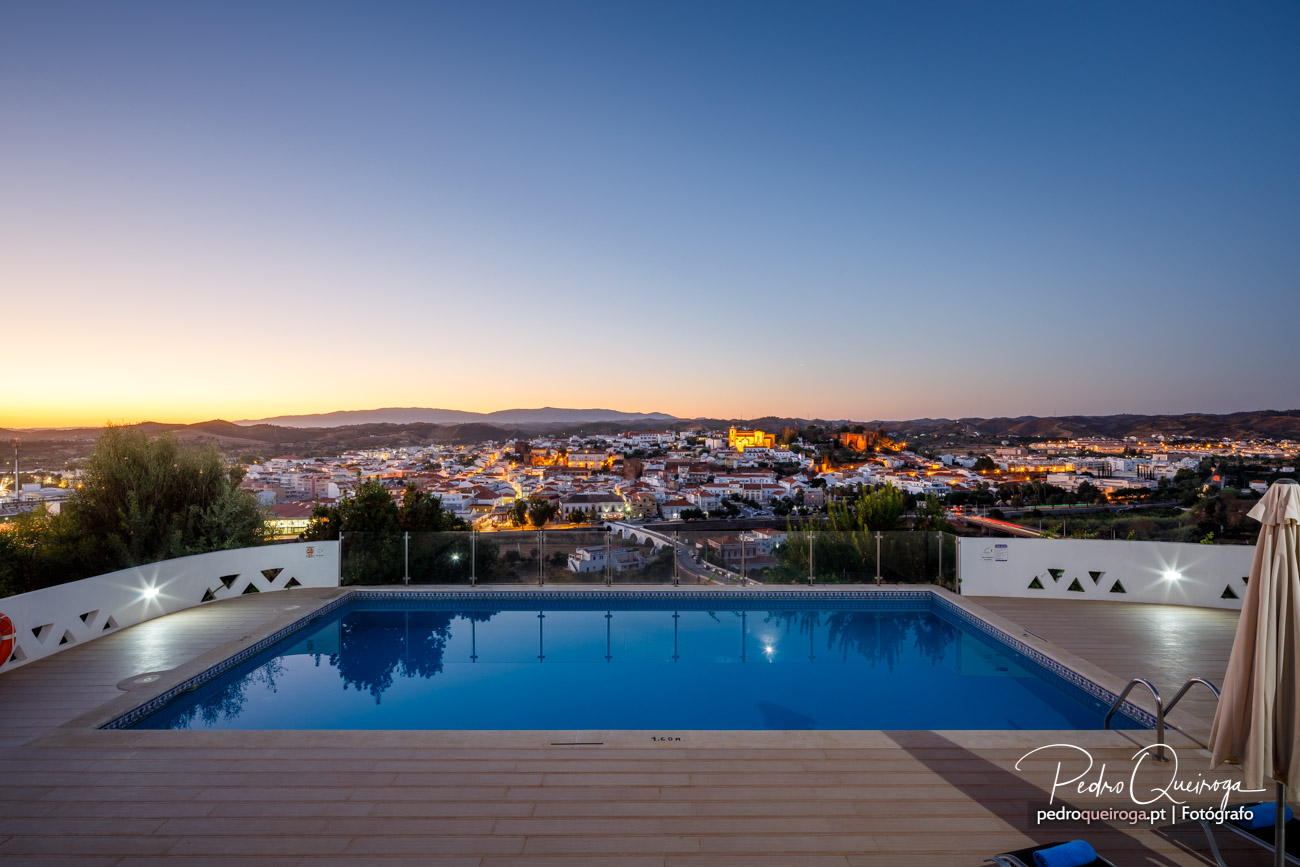 Piscina iluminada ao entardecer com vista panorâmica sobre a cidade e serra ao fundo, céu em tons dourados e azuis.