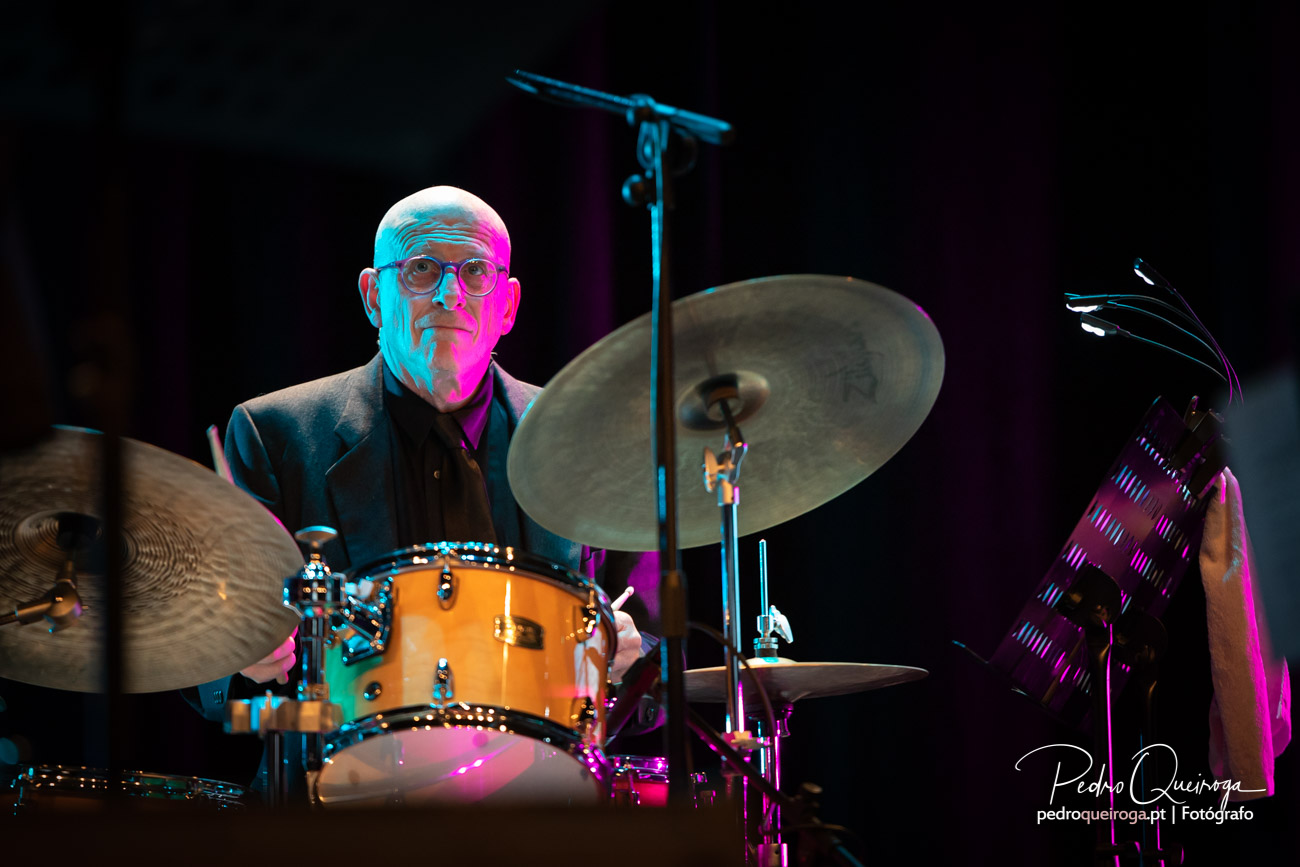 Baterista de óculos toca bateria laranja num palco com iluminação azul e rosa vibrante sobre fundo escuro