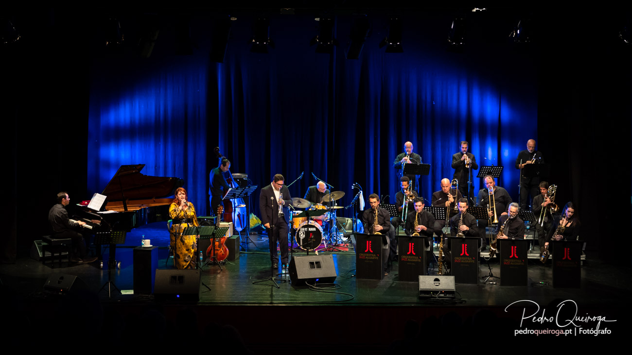 Orquestra de jazz atua em palco com cortina azul iluminada, pianista, vocalista e músicos com instrumentos de sopro