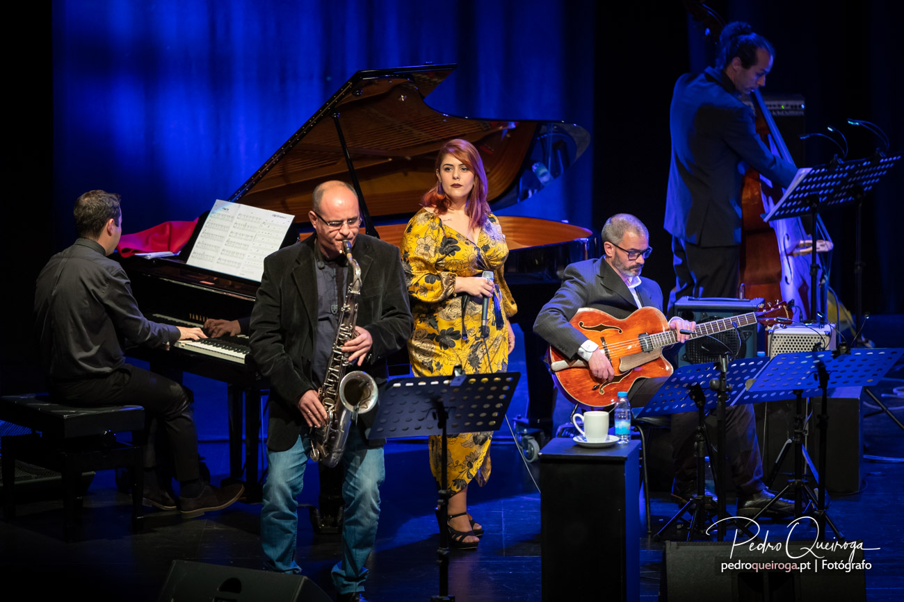 Quinteto de jazz em palco com piano, saxofone, vocalista de vestido amarelo, guitarrista e trompetista sob luz azul.