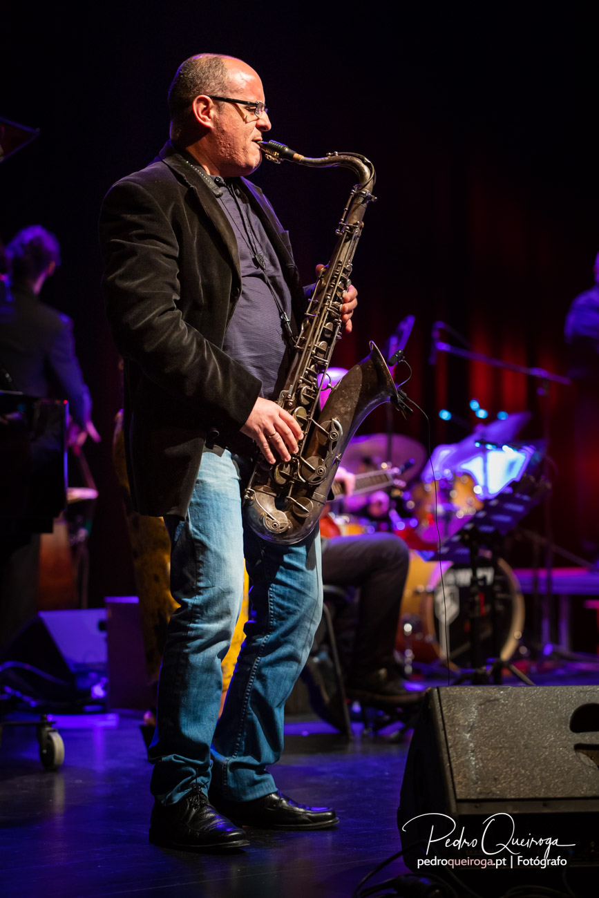 Saxofonista de óculos e blazer preto toca em palco iluminado por luz roxa com banda ao fundo