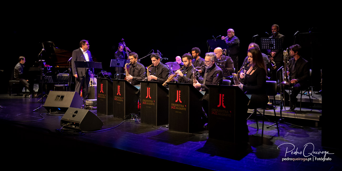 Orquestra de jazz alinhada no palco com arquitetura visual marcada por estantes e iluminação azul envolvente