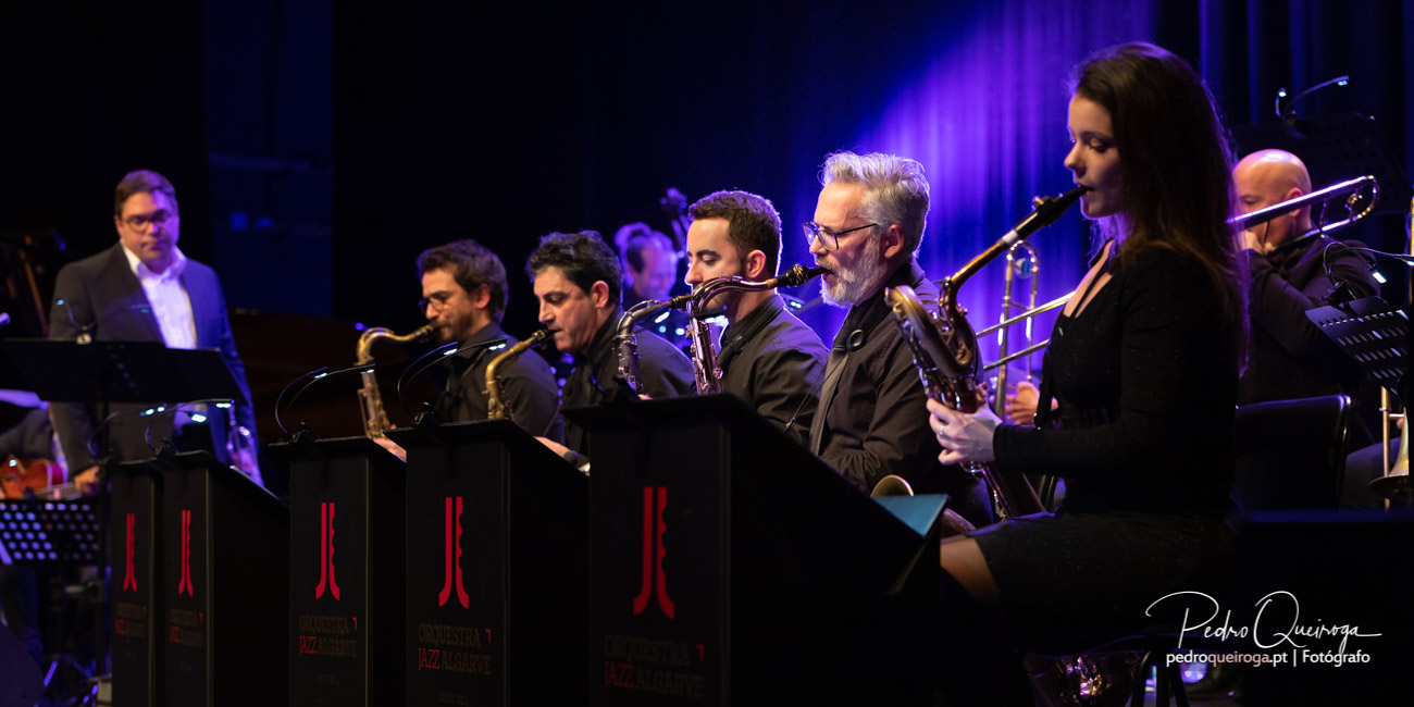 Orquestra de jazz em palco com saxofones e trompetes sob luz azul e violeta, numa arquitetura sonora envolvente