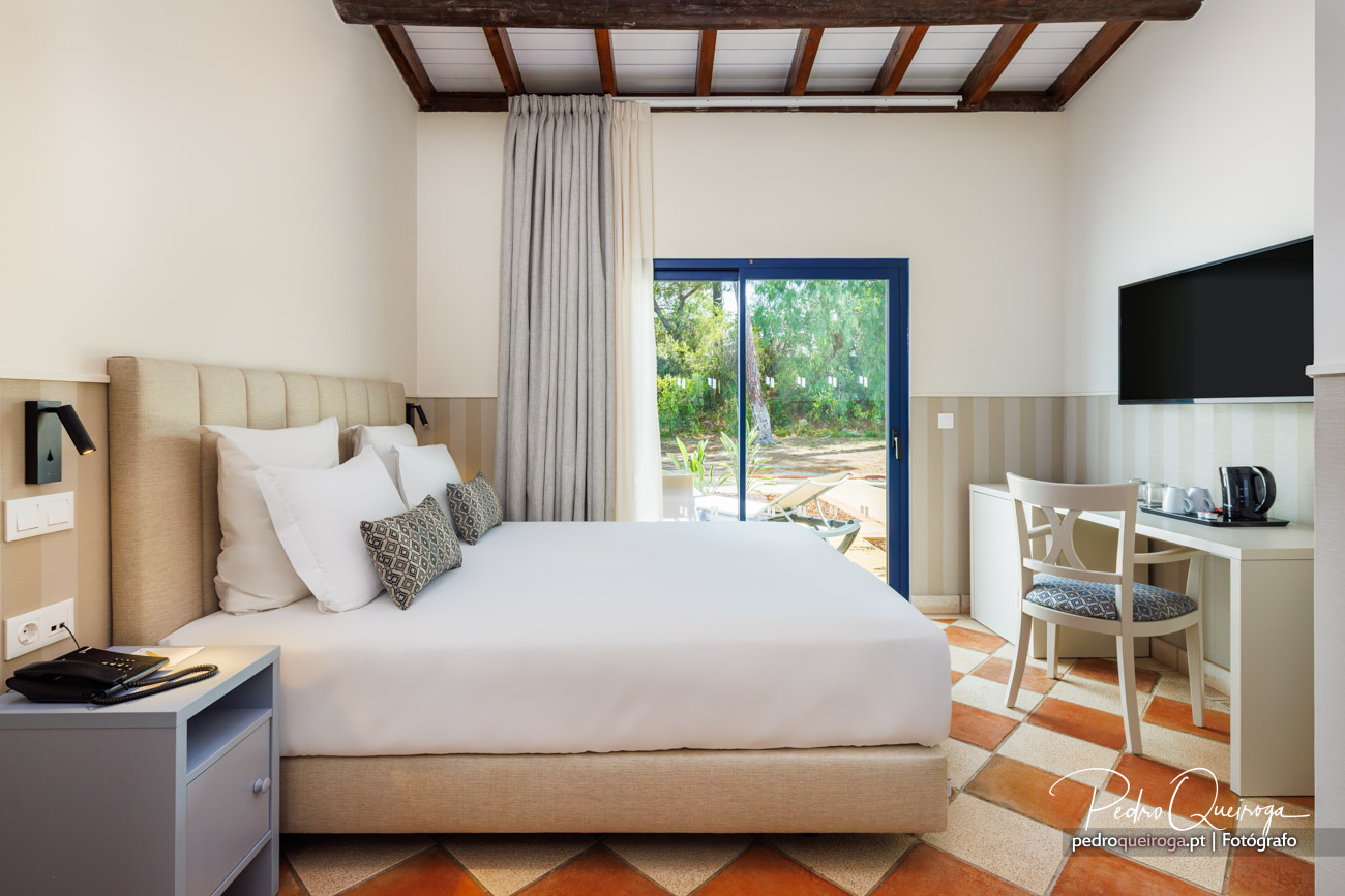 Quarto luminoso com cama bege, teto de madeira, janela com vista para jardim e pequena zona de trabalho