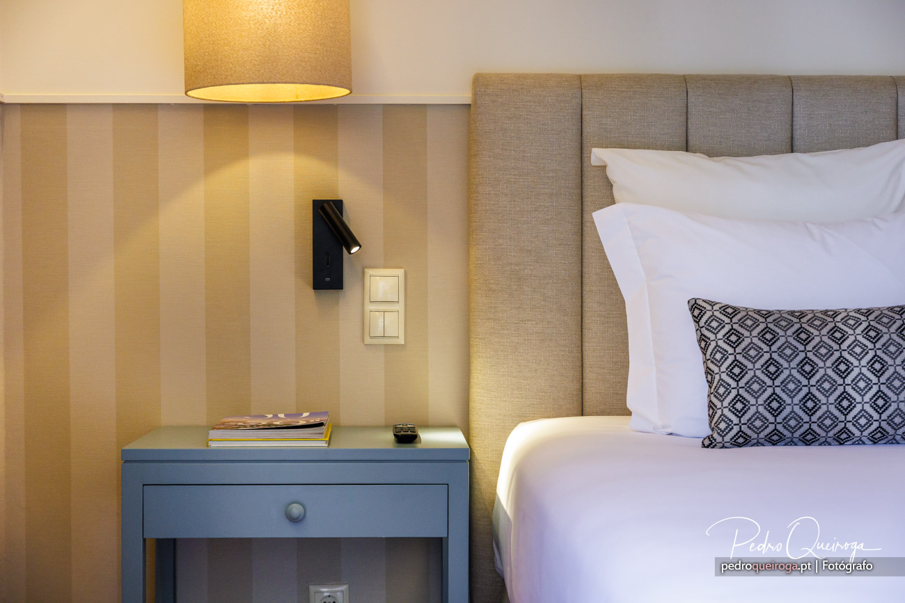 Quarto moderno com cabeceira estofada bege, mesinha azul, papel de parede às riscas e iluminação ambiente acolhedora.