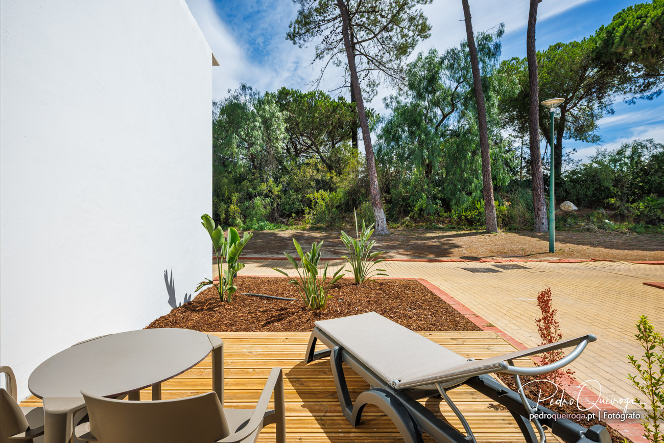 Terraço em deck de madeira com espreguiçadeiras e mesa, canteiro com plantas tropicais e pinhal em fundo.