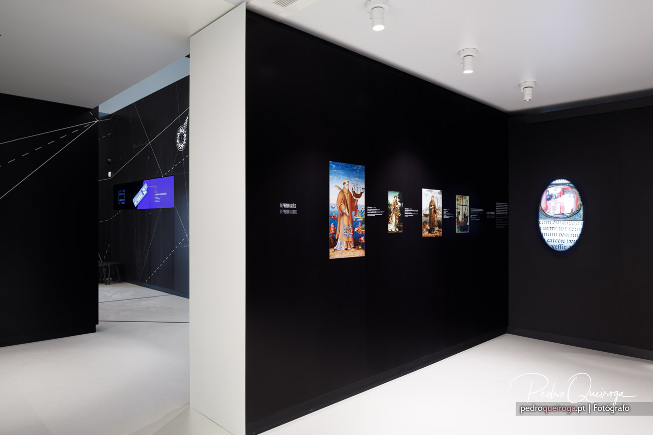Sala de exposição com paredes pretas, obras religiosas iluminadas e ecrã interativo ao fundo num espaço moderno e minimalista