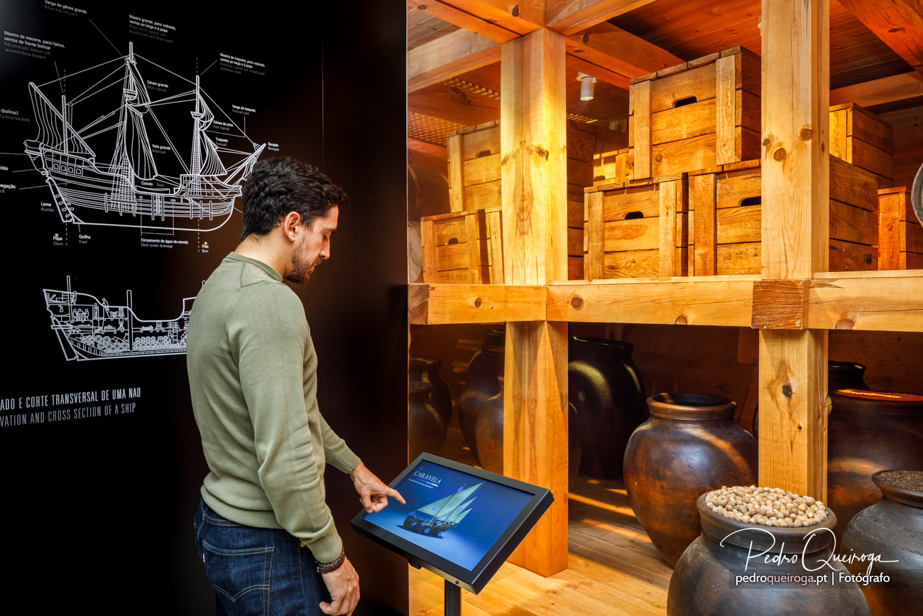 Homem a explorar ecrã interativo num museu marítimo, ao lado de estante com caixotes de madeira e jarras antigas
