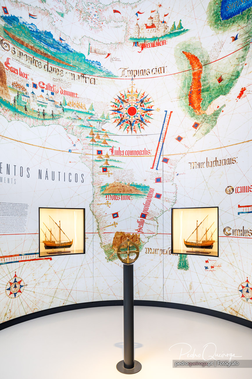 Mapa-múndi antigo ilustrado com caravelas, rosa dos ventos e caligrafia medieval em exposição museal interativa