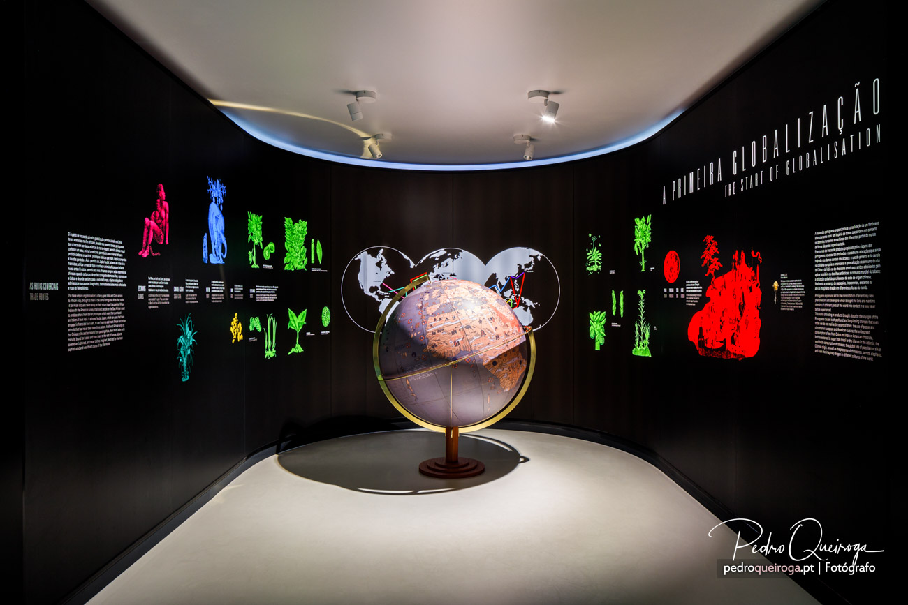 Globo terrestre iluminado ao centro de sala circular com paredes negras e figuras coloridas sobre globalização