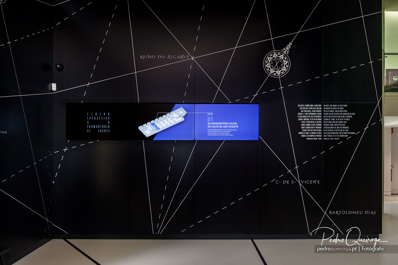 Parede preta com mapa geométrico do Algarve, ecrã azul ao centro exibindo maquete branca de promontório sagrado
