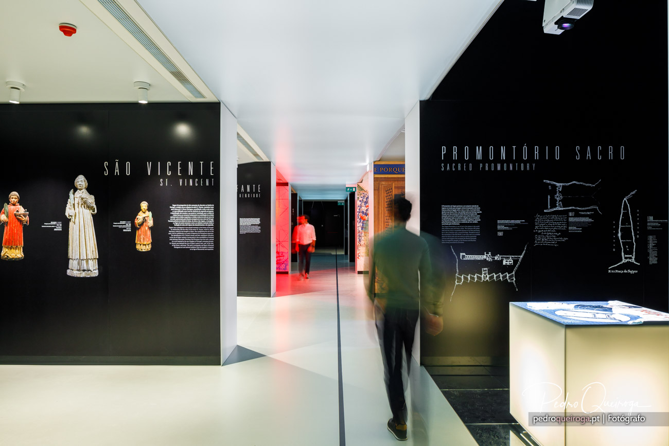 Visitante caminha entre salas expositivas com paredes pretas mostrando arte sacra e textos informativos num museu moderno